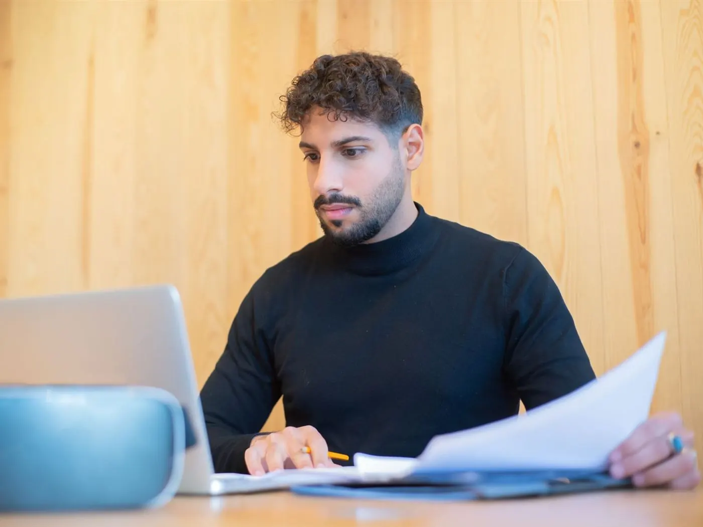 Homme en col roulé noir qui examine des documents devant son ordinateur portable