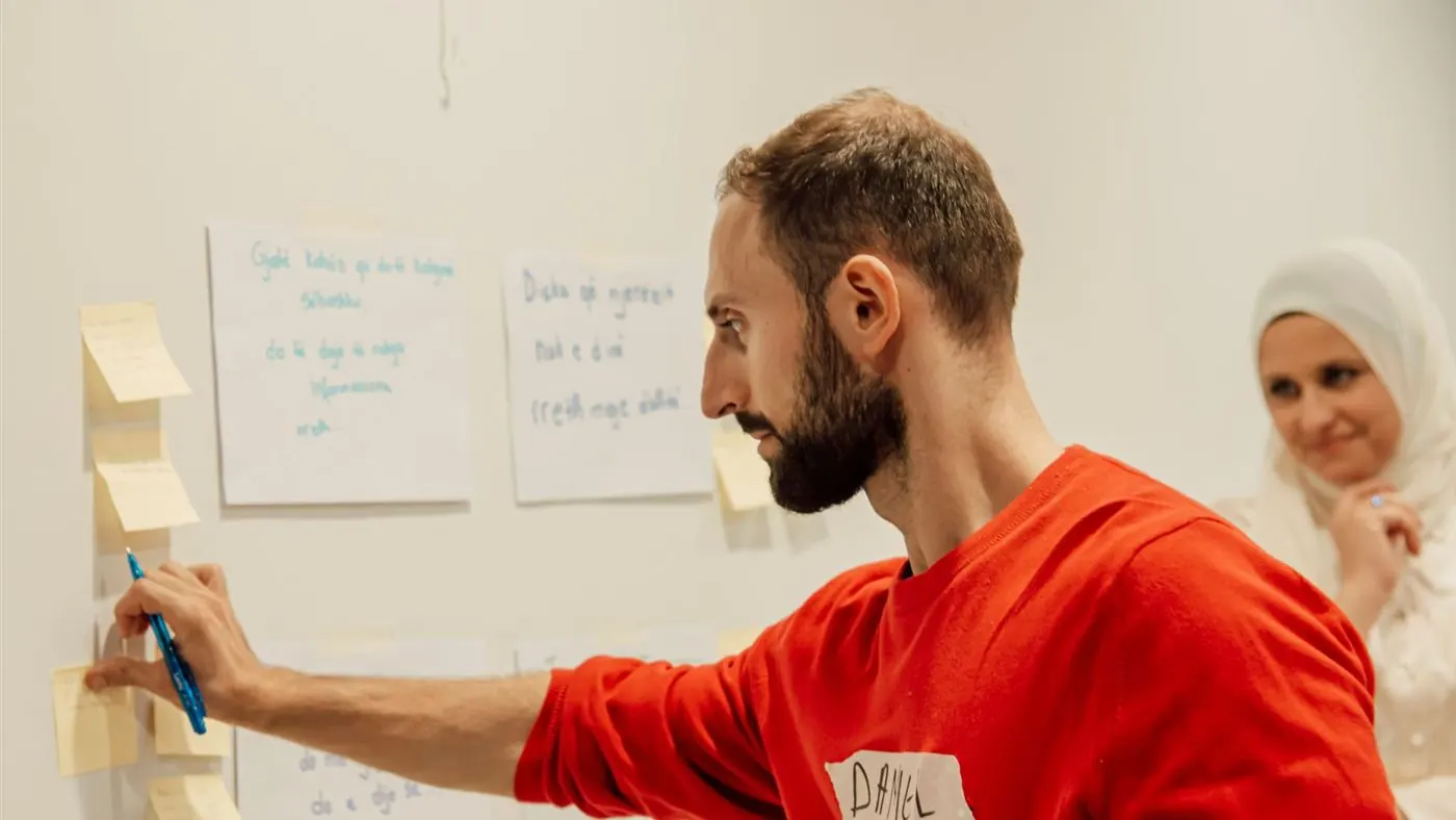 Homme qui colle une note adhésive sur un mur pendant un atelier de stratégie, une collègue sourit en arrière-plan
