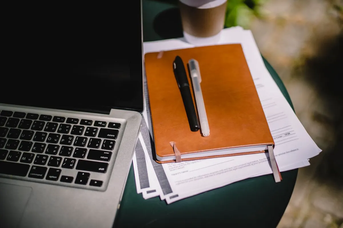 Carnet en cuir et stylos posés sur des documents, à côté d'un ordinateur portable, sur une table de jardin